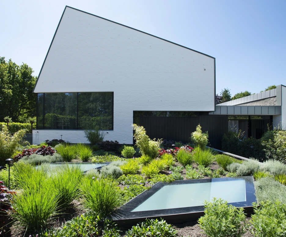 A modern white house with a distinctive angular roof, surrounded by a lush garden featuring various plants and small ponds. Bright blue sky is visible in the background.