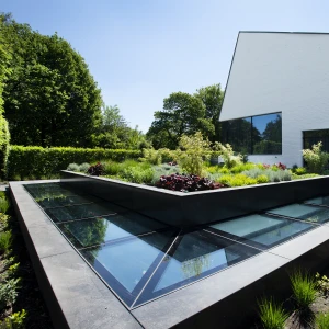 Jardín moderno con diseño geométrico en el centro, rodeado de vegetación variada y una casa blanca contemporánea al fondo. La estructura del jardín incluye paneles de vidrio que reflejan la luz del sol.