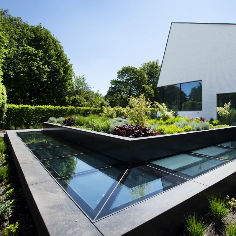 A modern building with a white facade sits beside a landscaped garden featuring a geometric arrangement of plants and glass features. The garden has varying greenery and colorful flowers, and the building boasts large windows reflecting the bright blue sky.