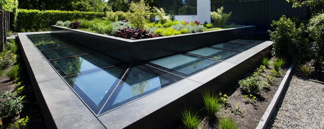 Jardín moderno con diseño geométrico en el centro, rodeado de vegetación variada y una casa blanca contemporánea al fondo. La estructura del jardín incluye paneles de vidrio que reflejan la luz del sol.