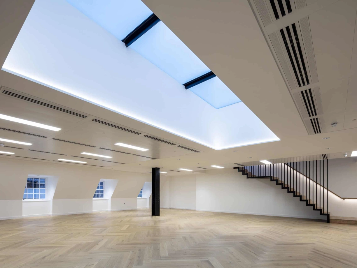 A large empty room with a staircase by the side and underneath a multipart skylight