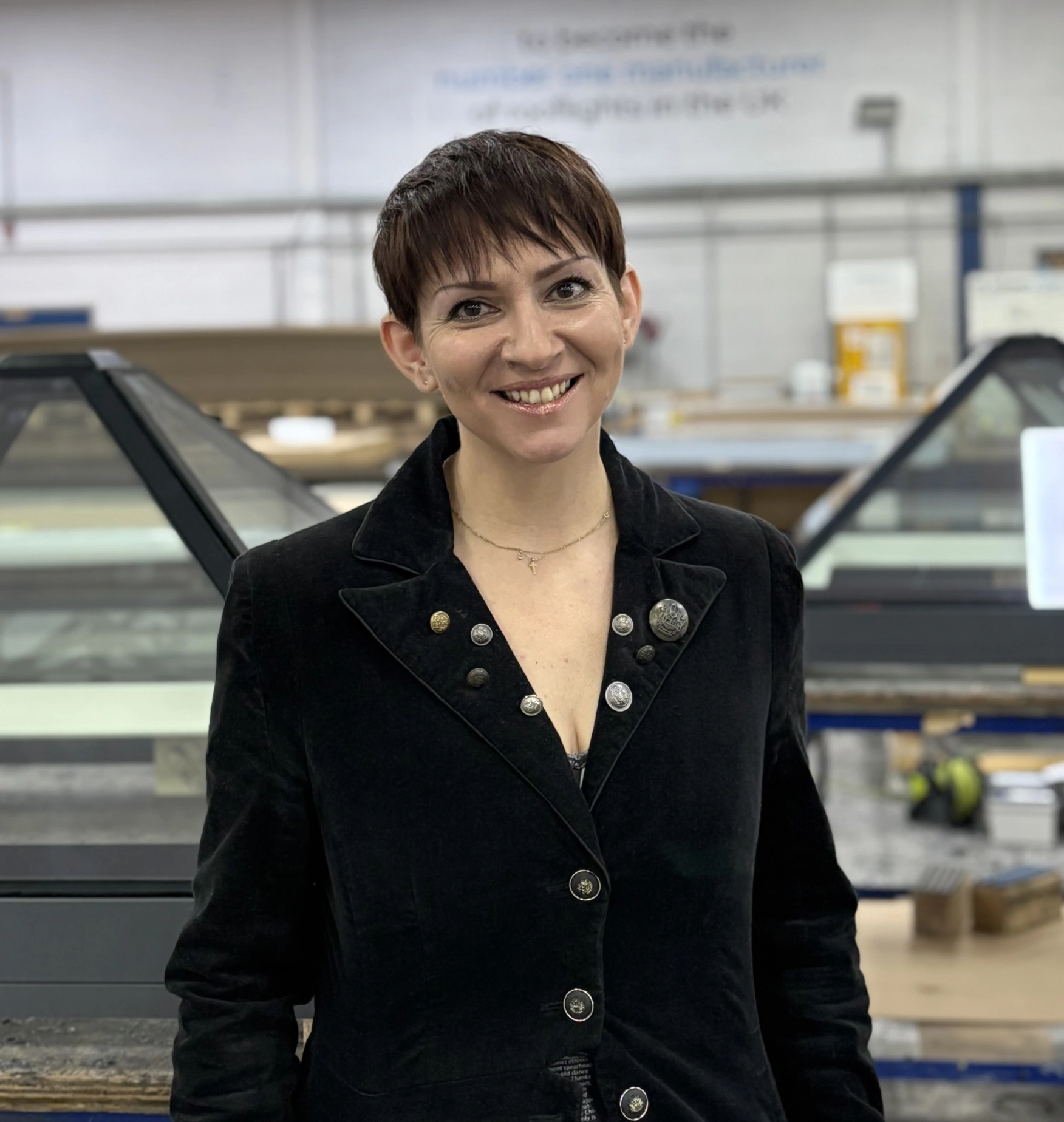 A woman with short hair smiles confidently while standing in a factory setting. She wears a black jacket with buttons and stands in front of glass panel structures. The background features a workspace with tools and equipment.