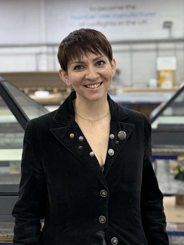 A woman with short hair smiles confidently while standing in a factory setting. She wears a black jacket with buttons and stands in front of glass panel structures. The background features a workspace with tools and equipment.