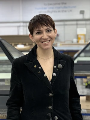 A woman with short hair smiles confidently while standing in a factory setting. She wears a black jacket with buttons and stands in front of glass panel structures. The background features a workspace with tools and equipment.