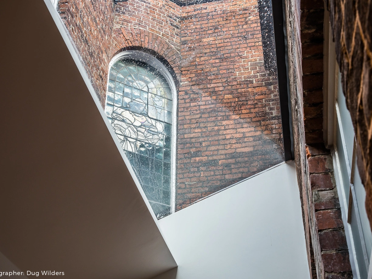 Light filters through a large stained glass window, illuminating the interior of a brick building. The contrast between the aged brick and modern white walls creates a unique architectural blend.