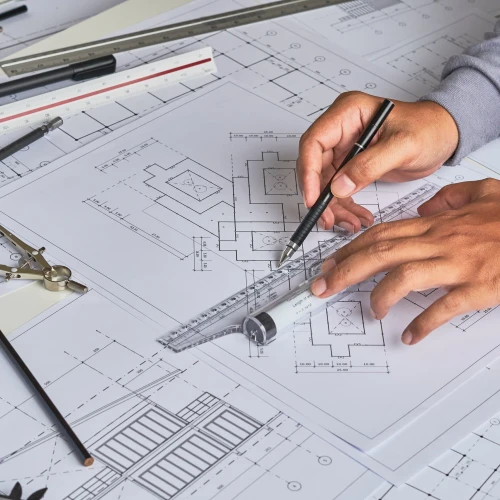 Des plans architecturaux étalés sur une table. Deux mains utilisent une règle et un stylo pour tracer des lignes sur les documents. Des outils de dessin, un compas et des crayons sont également visibles.