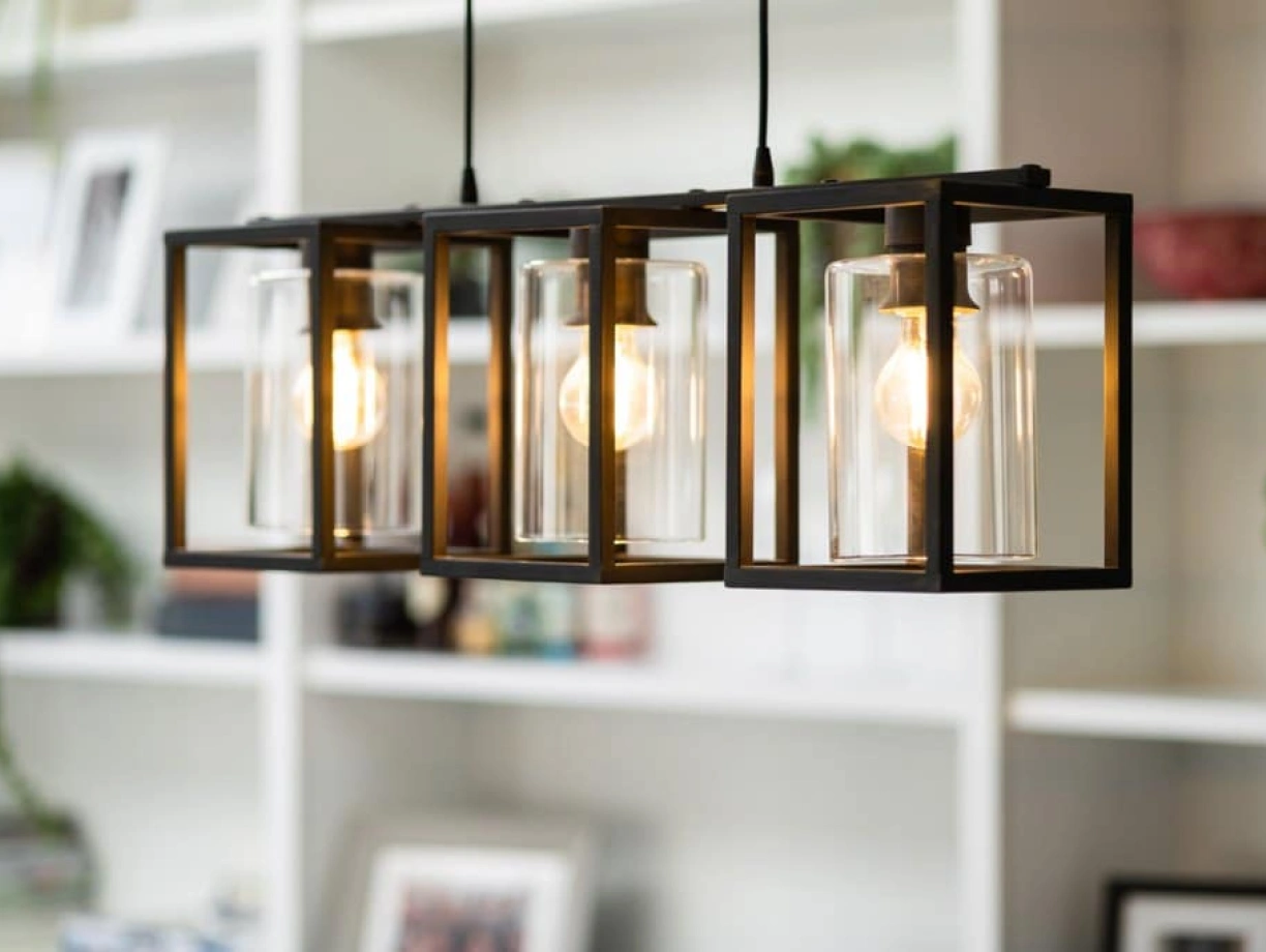 Three elegant pendant lights hang from a ceiling, each enclosed in a black metal frame with clear glass shades. The warm glow of the bulbs illuminates a softly decorated white shelf in the background, adorned with plants and decorative items.