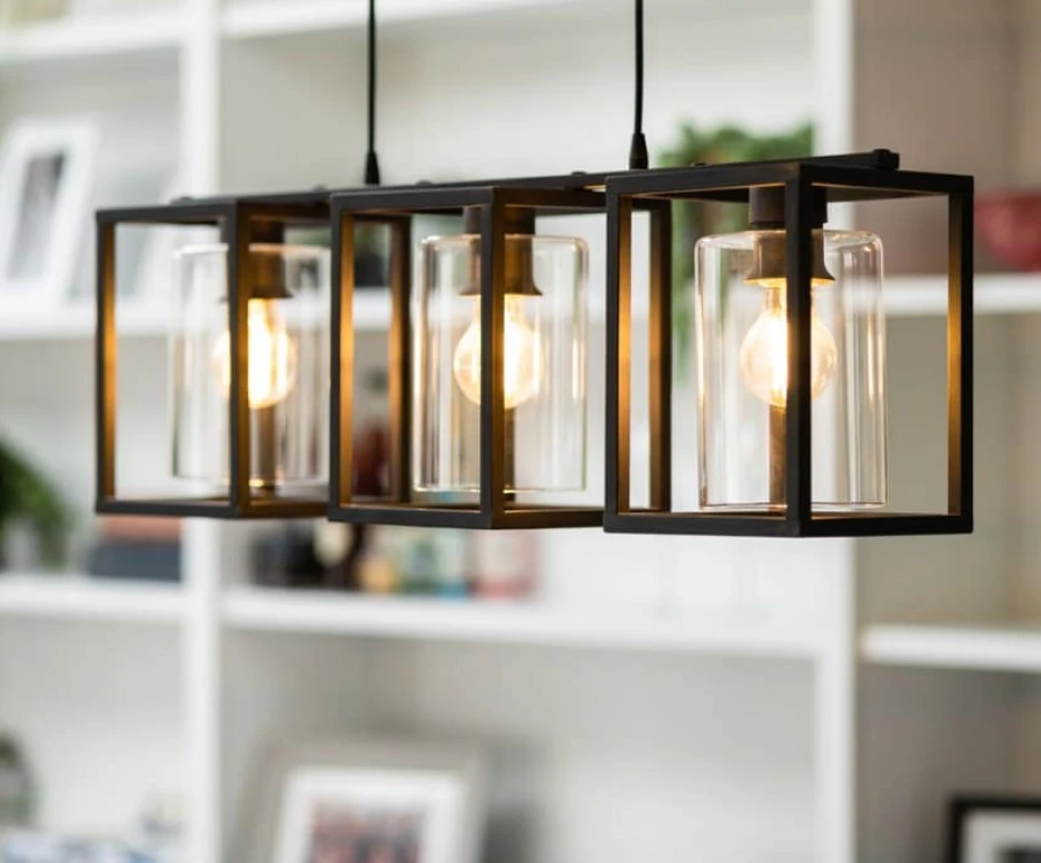 Three elegant pendant lights hang from a ceiling, each enclosed in a black metal frame with clear glass shades. The warm glow of the bulbs illuminates a softly decorated white shelf in the background, adorned with plants and decorative items.