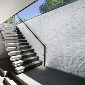 Modern staircase leading upwards, featuring sleek wooden steps and a glass railing. The walls are textured and white, with natural light coming in from above, illuminating the space.