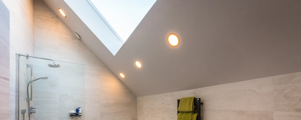 Modern bathroom featuring a freestanding bathtub, glass shower enclosure, and sleek white sink. The walls and floor are tiled in light stone, with an angled ceiling and skylight. A green towel hangs on a heated towel rack next to a bidet. Soft lighting highlights the space.