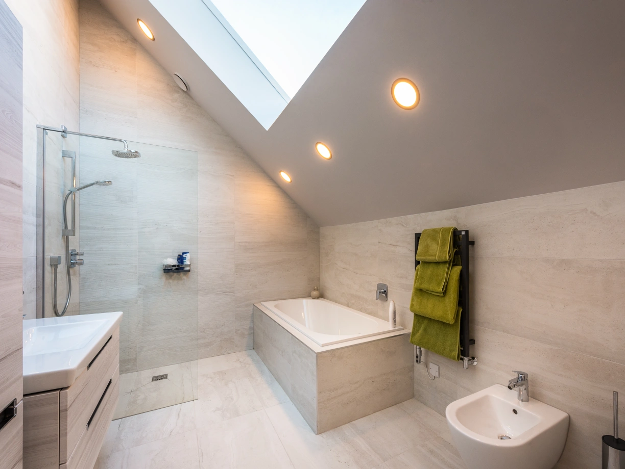 A modern bathroom featuring a freestanding bathtub, a glass shower stall, and a sleek vanity. The walls are light-colored with a minimalist design, and a green towel hangs on a heated towel rack. Natural light streams in from a skylight.