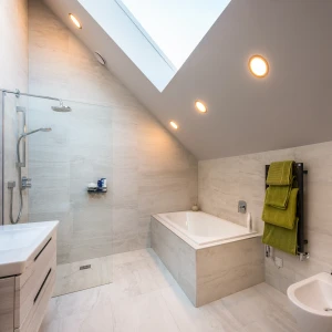 Modern bathroom featuring a freestanding bathtub, glass shower enclosure, and sleek white sink. The walls and floor are tiled in light stone, with an angled ceiling and skylight. A green towel hangs on a heated towel rack next to a bidet. Soft lighting highlights the space.
