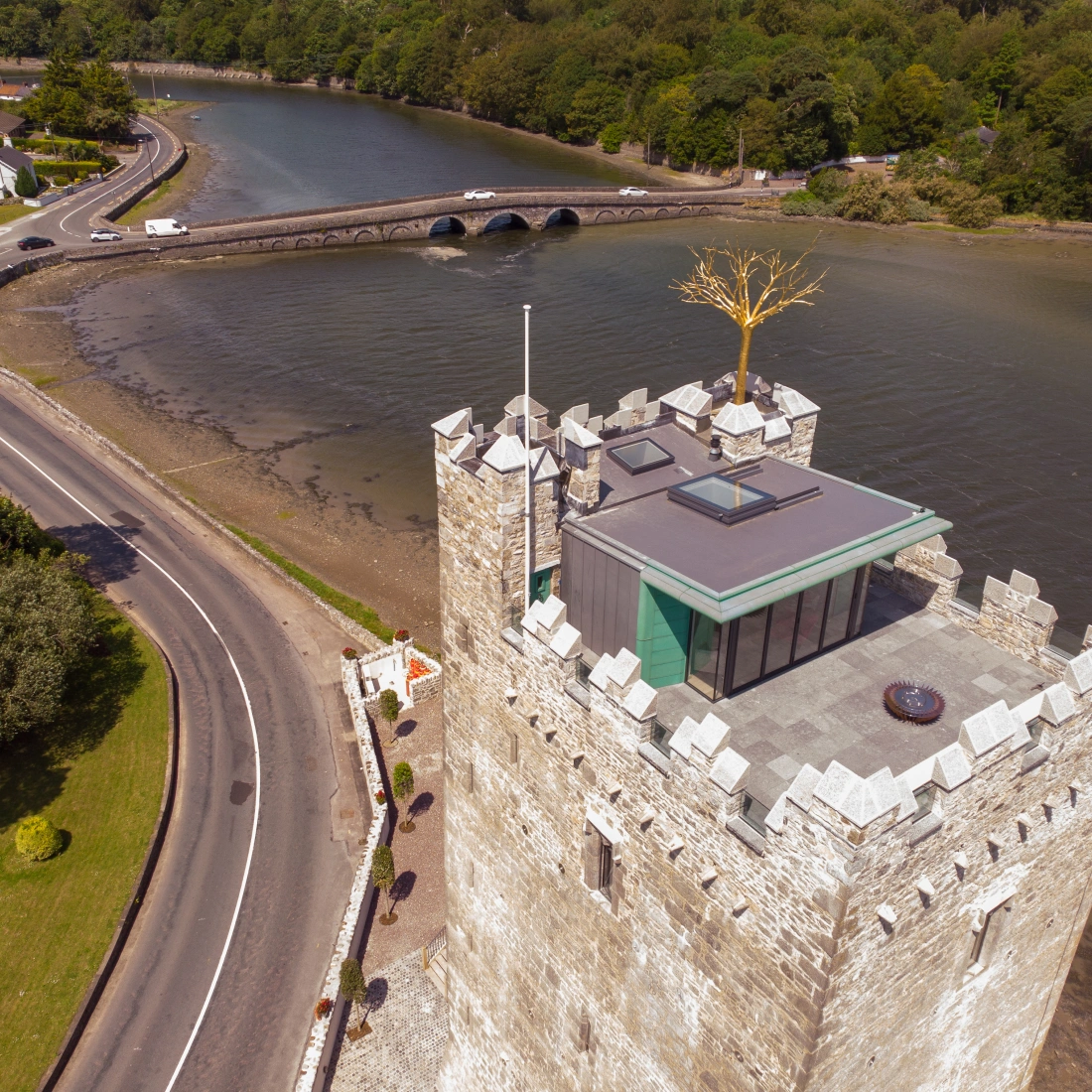 Aerial view of a stone castle tower beside a river, featuring a modern glass structure on top and a golden tree sculpture. A bridge crosses the river in the background, surrounded by lush greenery and a road.