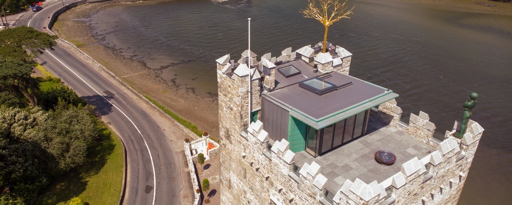 Aerial view of a stone tower with a modern glass structure on top, located near a river. Surrounding greenery and a road alongside the water are visible. A bridge crosses the river in the background.