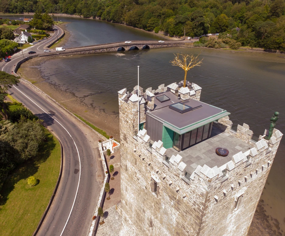 Aerial view of a stone tower with a modern glass structure on top, located near a river. Surrounding greenery and a road alongside the water are visible. A bridge crosses the river in the background.