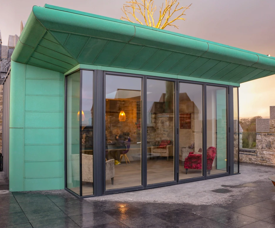 A modern green building with large glass windows, situated on a stone structure. The roof has a unique angular design, and there are stylish furnishings visible inside. The surrounding area features a stone wall, and the sky is cloudy.