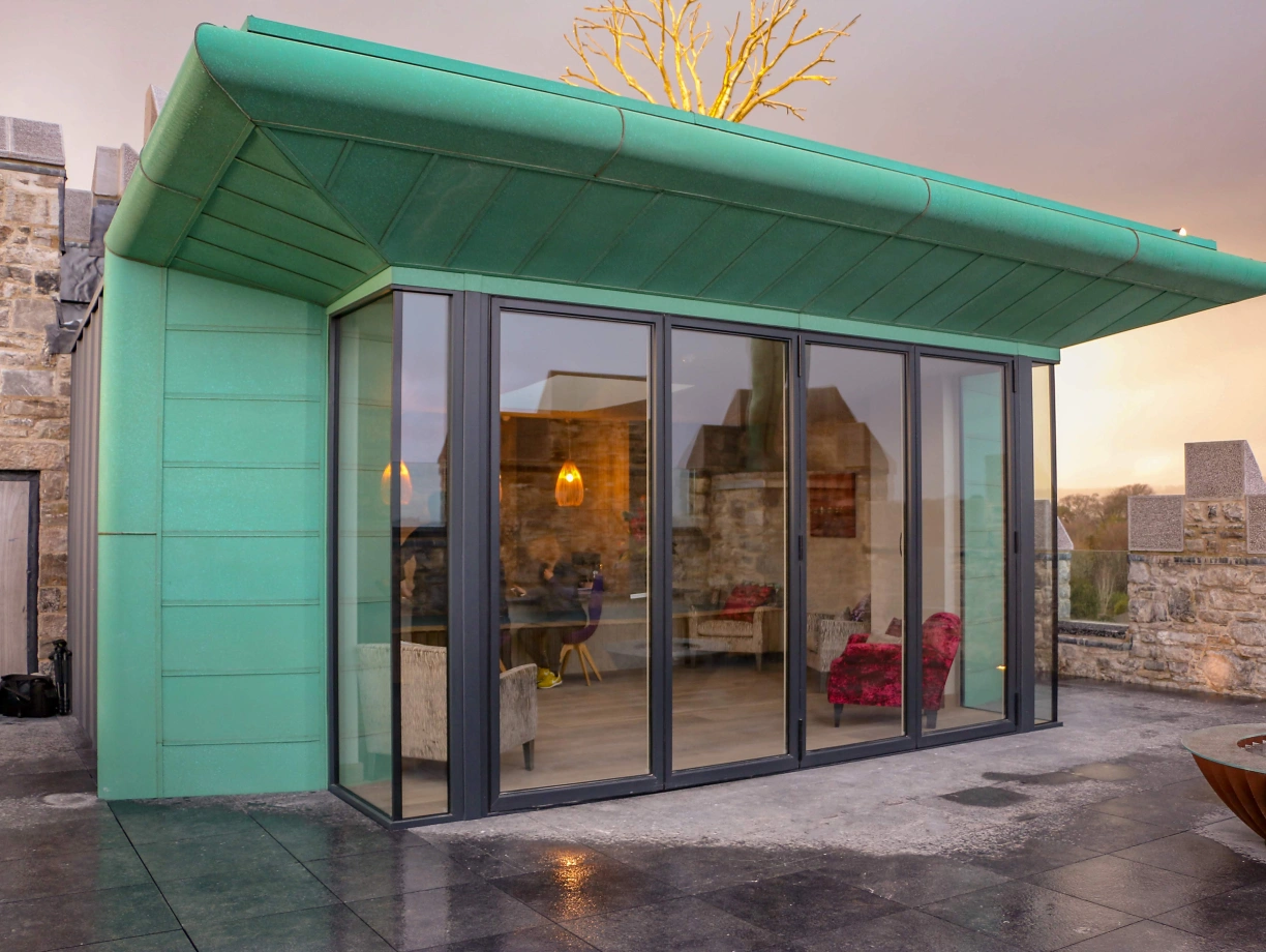 A modern green building with large glass windows, situated on a stone structure. The roof has a unique angular design, and there are stylish furnishings visible inside. The surrounding area features a stone wall, and the sky is cloudy.