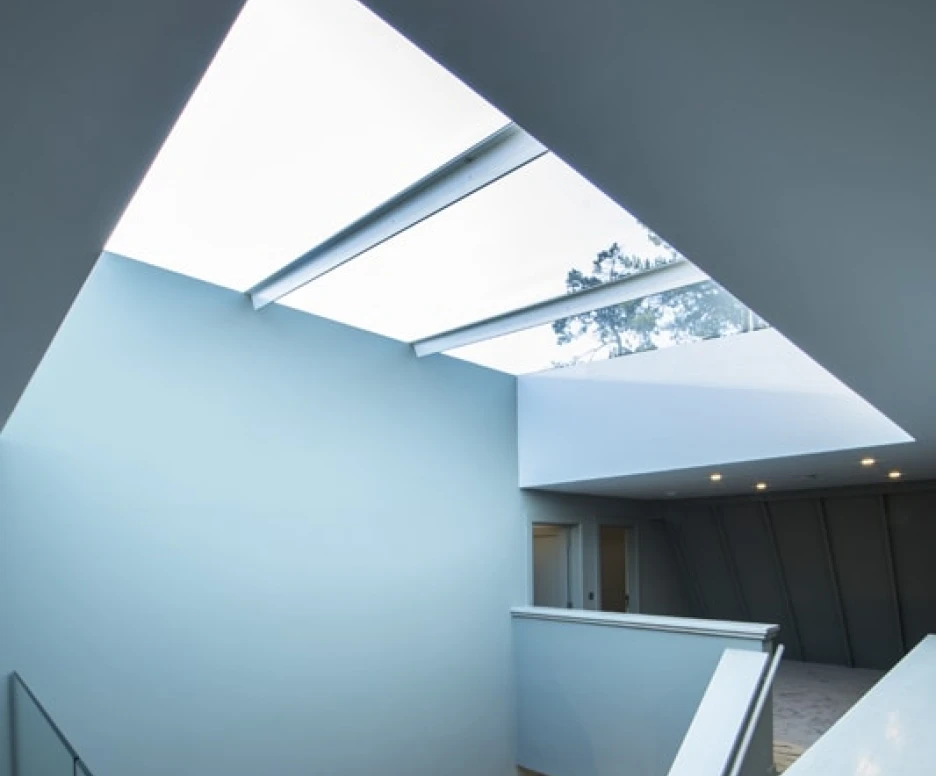 A modern staircase with a glass railing, leading to an upper level. Natural light streams through a skylight above, illuminating the pale blue walls. The space is stylish and minimalist, with soft lighting fixtures enhancing the ambiance.