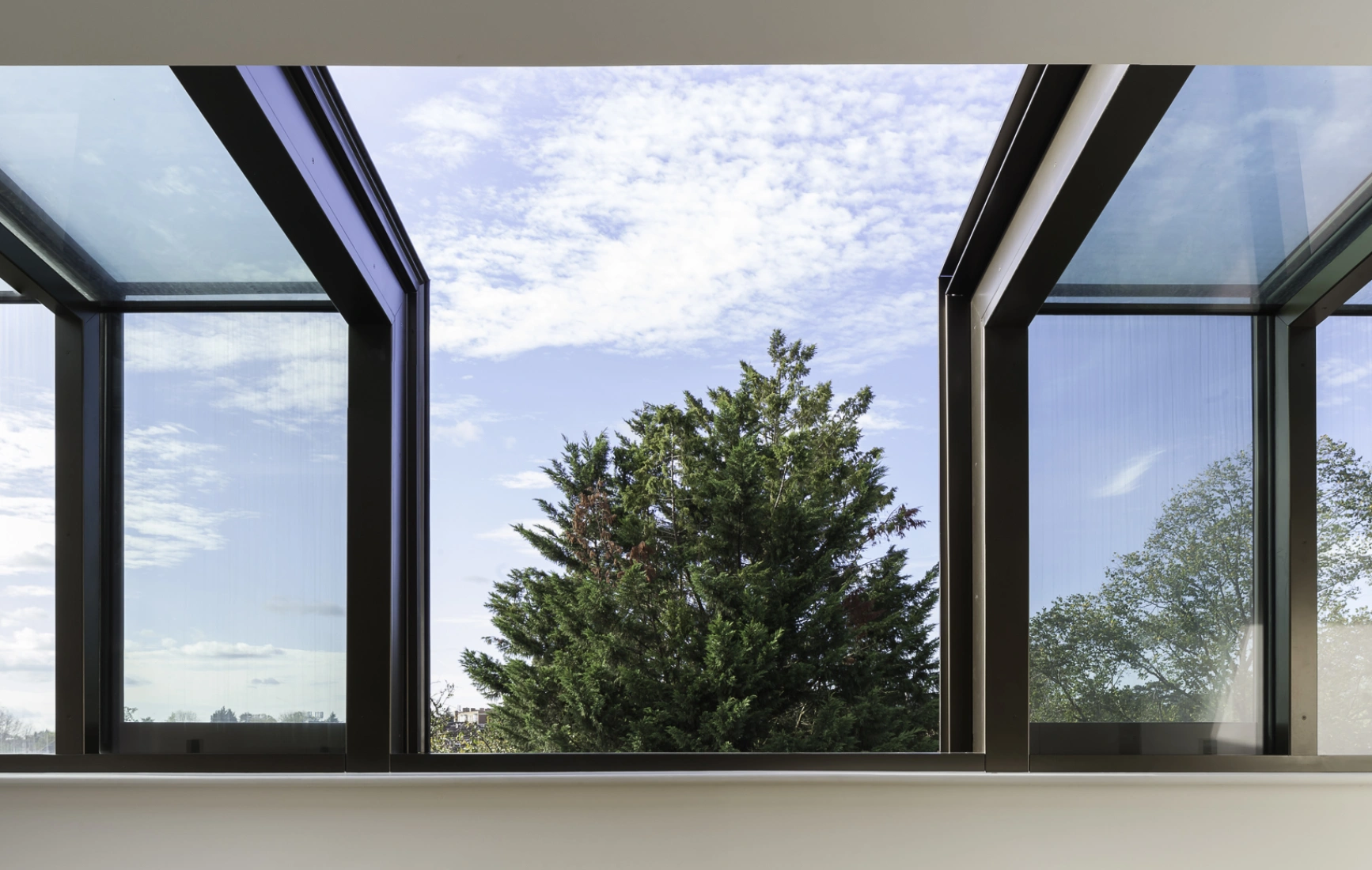 Vue intérieure d’une pièce avec de grandes fenêtres ouvertes. Ciel nuageux et un arbre vert visible à l’extérieur. Sol en bois clair et mobilier minimaliste.