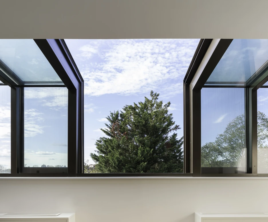 Vue intérieure d’une pièce avec de grandes fenêtres ouvertes. Ciel nuageux et un arbre vert visible à l’extérieur. Sol en bois clair et mobilier minimaliste.