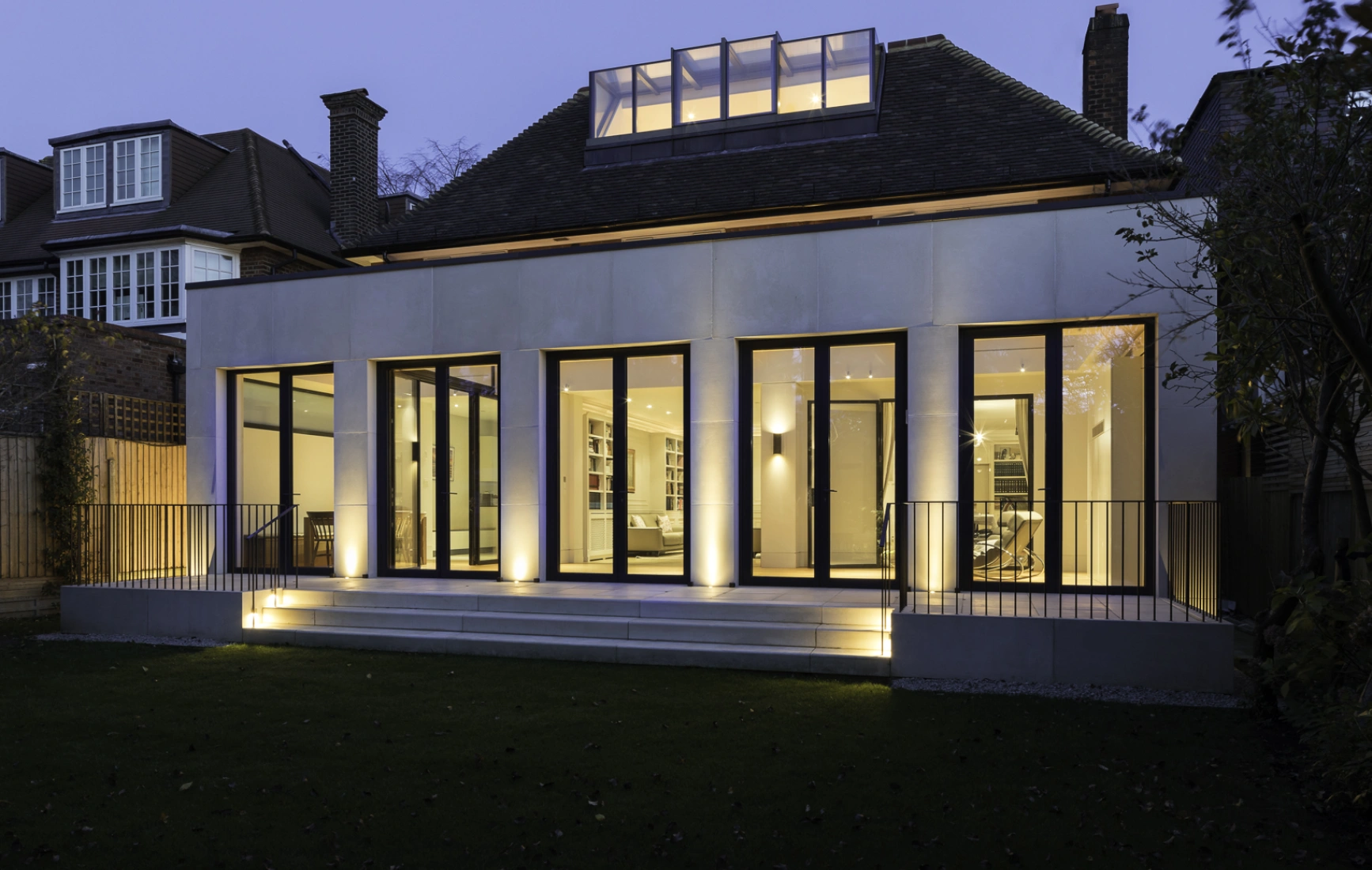 A modern house at dusk, featuring large glass windows that illuminate the interior. The structure is surrounded by a green lawn and has a wooden fence in the background. The roof has a skylight, adding to its contemporary design.