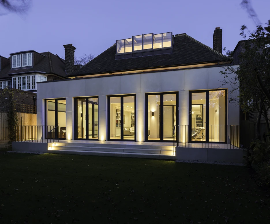 Casa moderna al anochecer, con fachada de grandes ventanales que iluminan el interior. El jardín está bien cuidado y la iluminación exterior resalta la arquitectura.