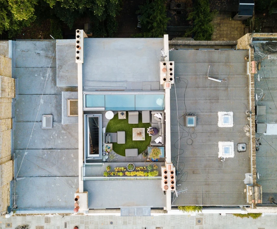 Aerial view of a modern rooftop terrace featuring a green lawn, seating area with outdoor furniture, and planters. Surrounding buildings and a tree-lined street are visible below.
