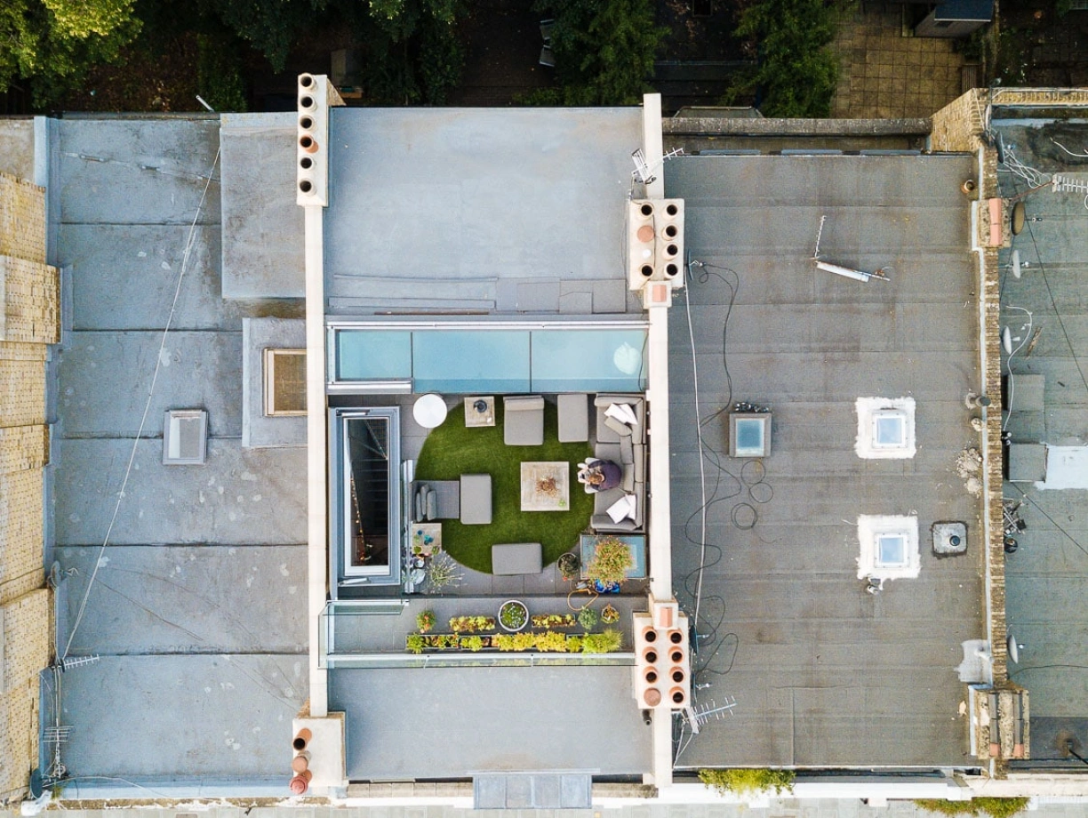 Aerial view of a modern rooftop garden featuring green grass, outdoor seating, and planters. Surrounding buildings and trees are visible, along with parked cars on the street below.