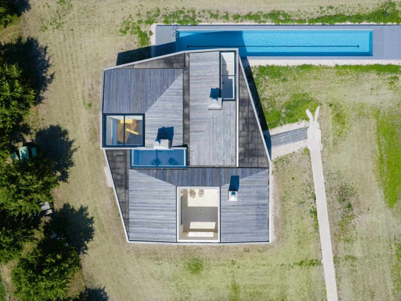 Aerial view of a modern, angular house with a flat roof and wooden deck. A long swimming pool runs alongside the property, which is surrounded by grassy fields and a winding pathway.