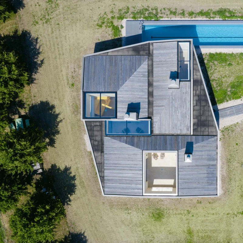 Aerial view of a modern, angular house surrounded by grassy land, featuring a rectangular swimming pool alongside. The house has a flat roof with patio areas and large windows. A pathway leads to the structure, enhancing the contemporary design.