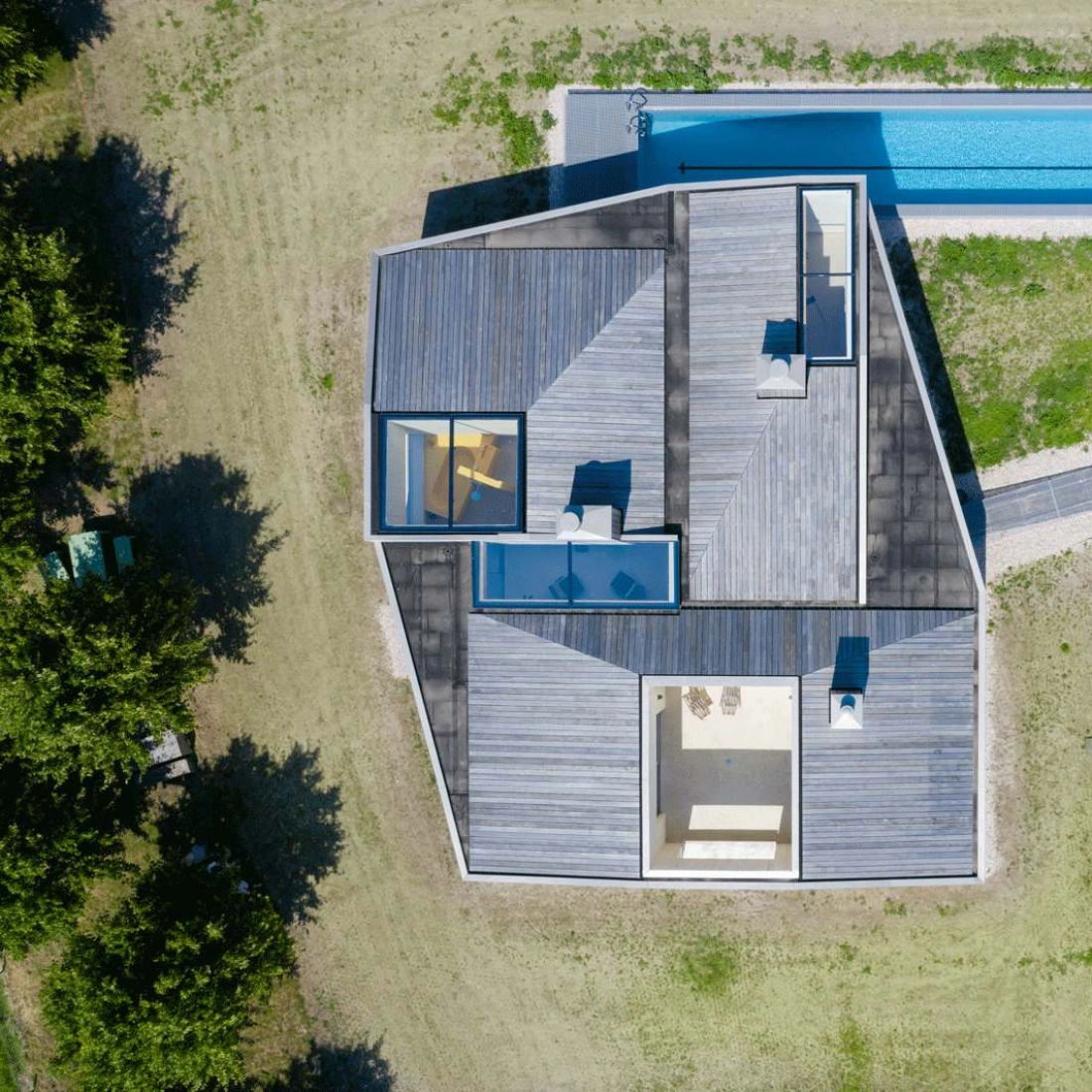 Vista aérea de una casa moderna con un diseño angular. La estructura tiene un patio y una piscina rectangular al lado. Rodeada de césped verde, la casa cuenta con áreas al aire libre y grandes ventanas.