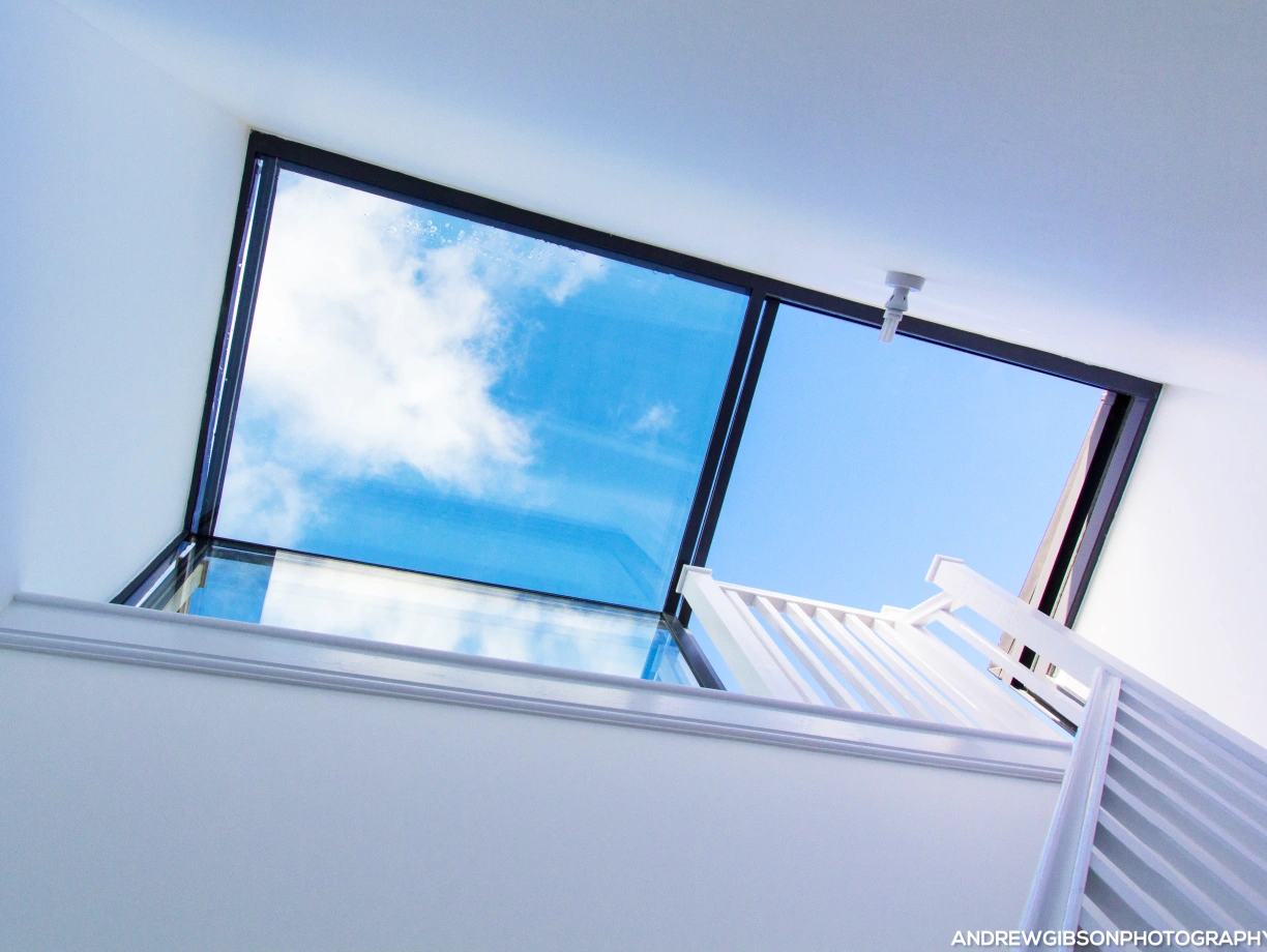 A modern stairwell with a glass skylight showing blue sky and clouds above. White walls and a railing line the staircase leading up.
