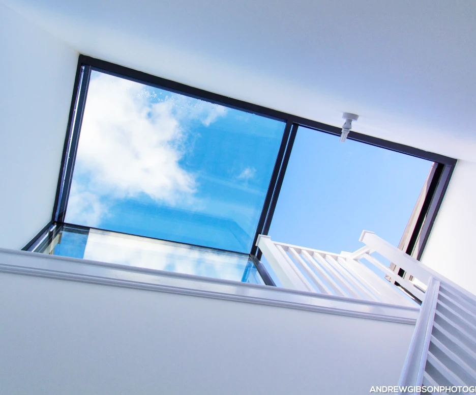 A modern stairwell with a glass skylight showing blue sky and clouds above. White walls and a railing line the staircase leading up.
