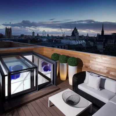 A stylish rooftop terrace features a modern seating area with a black wicker sofa and white cushions, alongside a sleek glass coffee table. A transparent hot tub is set within a black enclosure, with potted plants enhancing the decor. The backdrop showcases a breathtaking city skyline under a twilight sky.
