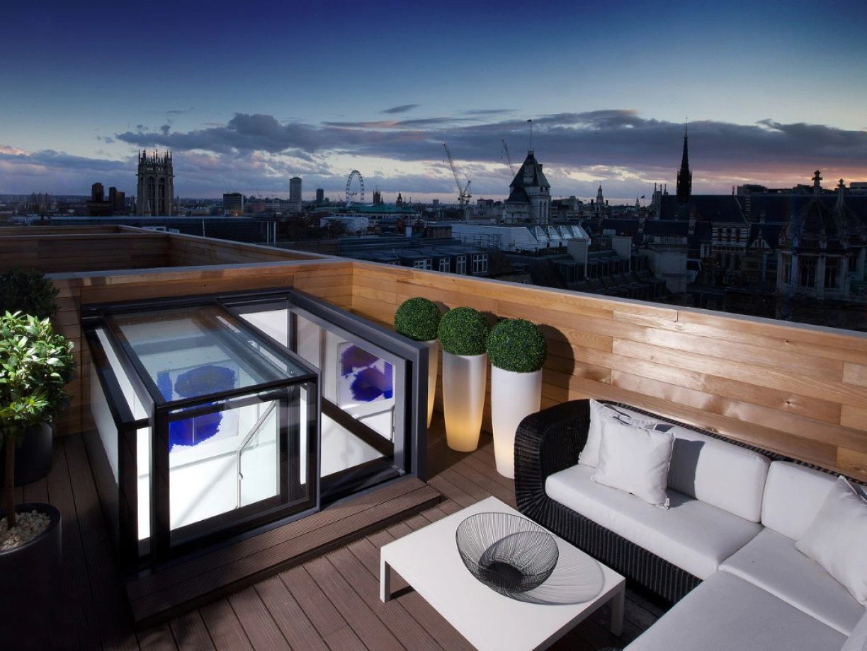 A stylish rooftop terrace features a modern seating area with a black wicker sofa and white cushions, alongside a sleek glass coffee table. A transparent hot tub is set within a black enclosure, with potted plants enhancing the decor. The backdrop showcases a breathtaking city skyline under a twilight sky.