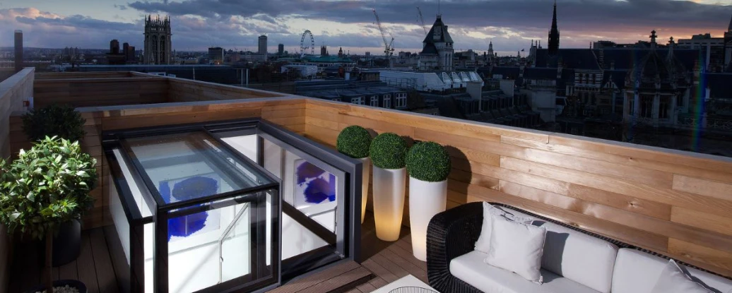 A stylish rooftop terrace features a modern seating area with a black wicker sofa and white cushions, alongside a sleek glass coffee table. A transparent hot tub is set within a black enclosure, with potted plants enhancing the decor. The backdrop showcases a breathtaking city skyline under a twilight sky.