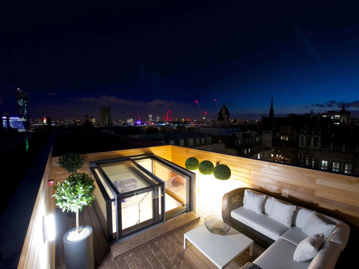 A rooftop terrace with modern furniture overlooking a city skyline at night. The scene features illuminated buildings, a Ferris wheel, and a gradient sky transitioning from blue to dark. Decorative plants add a touch of greenery to the setting.