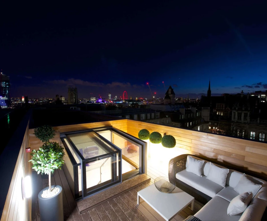 A rooftop terrace with modern furniture overlooking a city skyline at night. The scene features illuminated buildings, a Ferris wheel, and a gradient sky transitioning from blue to dark. Decorative plants add a touch of greenery to the setting.