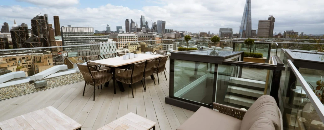 A spacious rooftop terrace featuring outdoor seating and dining arrangements, with a panoramic view of a city skyline that includes modern skyscrapers under a partly cloudy sky.