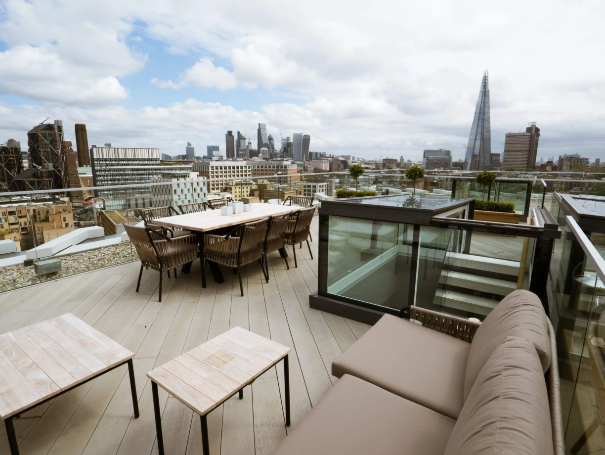 A spacious rooftop terrace featuring outdoor seating and dining arrangements, with a panoramic view of a city skyline that includes modern skyscrapers under a partly cloudy sky.