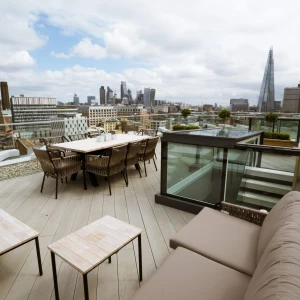 A spacious rooftop terrace featuring outdoor seating and dining arrangements, with a panoramic view of a city skyline that includes modern skyscrapers under a partly cloudy sky.