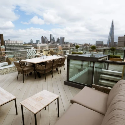 A spacious rooftop terrace featuring outdoor seating and dining arrangements, with a panoramic view of a city skyline that includes modern skyscrapers under a partly cloudy sky.