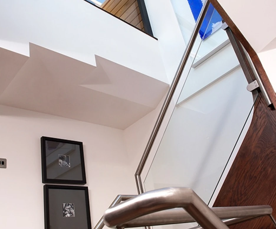 Modern staircase with a sleek metal railing ascending towards a skylight, showcasing a blue painting on the wall. Natural light brightens the interior, highlighting a minimalist design with framed art on the wall.