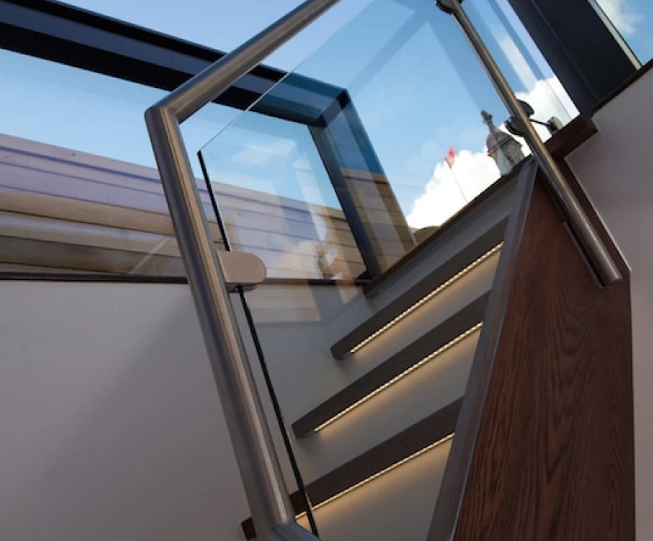 A modern staircase with wooden steps and a glass railing leads upward, illuminated by soft lighting. The sky is visible through a large skylight above.