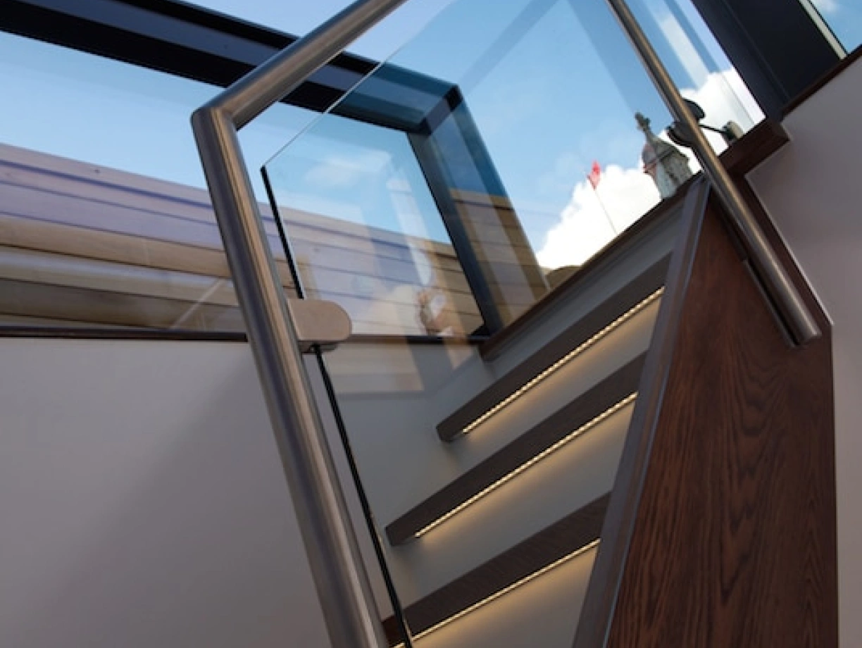 A modern staircase with wooden steps and a glass railing leads upward, illuminated by soft lighting. The sky is visible through a large skylight above.