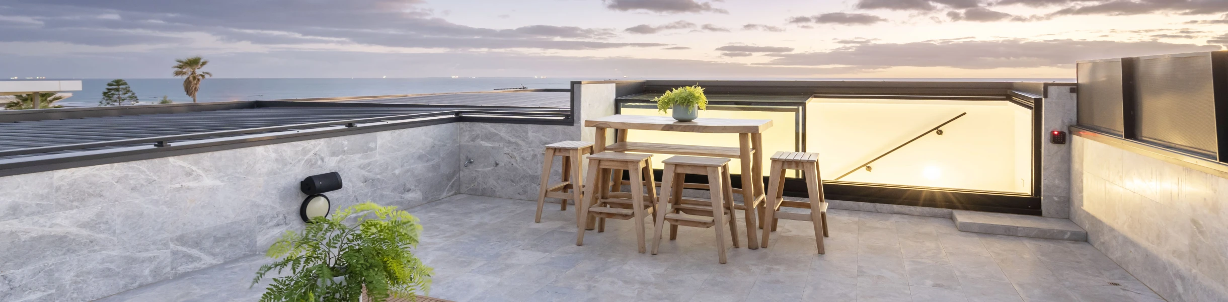 Terrasse spacieuse avec une table et des chaises en bois, décorée d'une plante en pot. Vue sur un ciel coloré au coucher de soleil avec des nuages. Un palmier est visible au loin.
