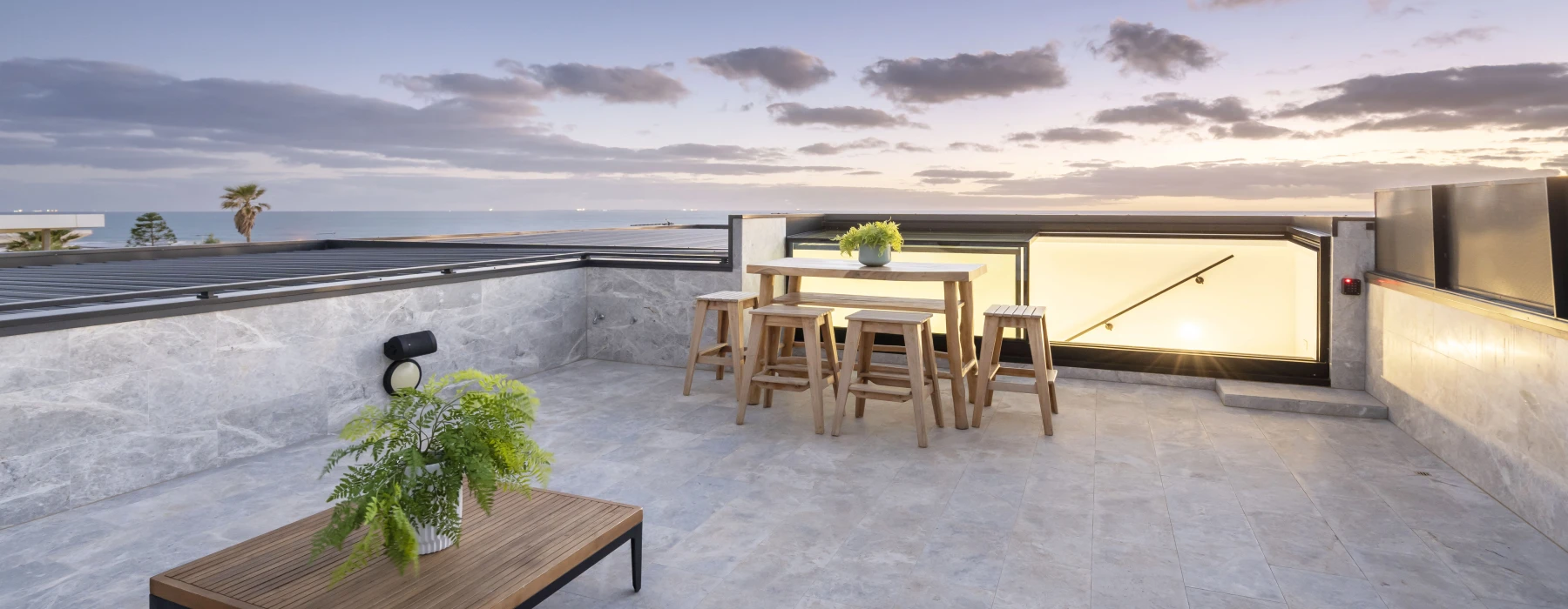 Terraza moderna con mesas y sillas de madera, decorada con plantas. Se observa un atardecer con nubes y palmeras en el horizonte.