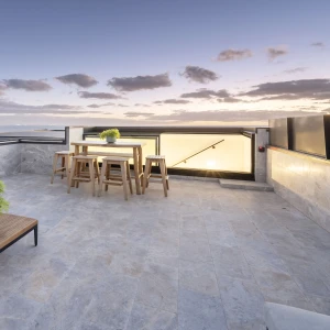 A modern rooftop terrace with a wooden table and stools, adorned with a small potted plant. The space features gray stone flooring and has a stunning view of the horizon under a twilight sky filled with clouds.