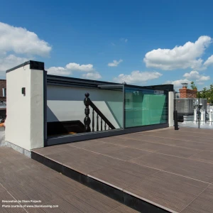 A rooftop space featuring wooden decking, glass railings, and a staircase. Bright blue sky with a few fluffy clouds is visible in the background.