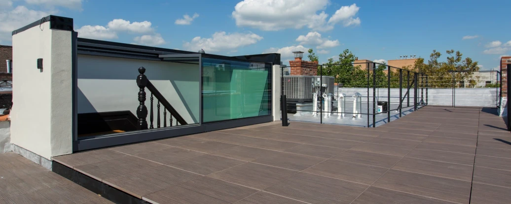 A rooftop space featuring wooden decking, glass railings, and a staircase. Bright blue sky with a few fluffy clouds is visible in the background.